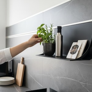 Può includere: Mensola in metallo nero con una pianta in vaso, una bottiglia in acciaio inossidabile e due piccoli libri. Una mano raggiunge l'erba. La mensola è montata su una parete piastrellata grigia, suggerendo una cucina.