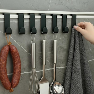 Könnte beinhalten: Ein Küchenorganisationssystem mit einer Metallschiene und schwarzen Haken. Eine Wurst, Küchenutensilien und ein graues Handtuch hängen an den Haken. Die Gegenstände werden vor einer grau gefliesten Wand präsentiert.