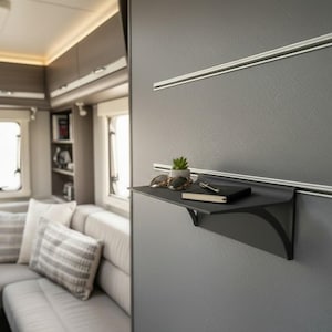Könnte beinhalten: Schwarzes Wandregal mit einer kleinen Pflanze, Sonnenbrille, Stift und Notizbuch. Das Regal ist an einer grauen Wand mit einer silbernen Schiene darüber befestigt. Der Innenraum eines Wohnmobils ist im Hintergrund zu sehen.