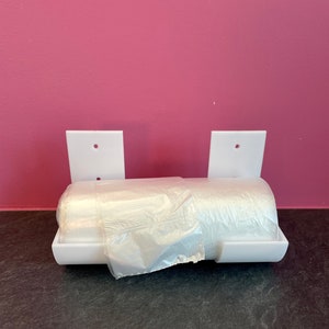 Può includere: Distributore da parete in plastica bianca per contenere un rotolo di sacchetti di plastica. Il distributore ha due staffe bianche con fori per il montaggio e una base in plastica bianca con una fessura per il rotolo di sacchetti. Un rotolo di sacchetti di plastica trasparente è parzialmente inserito nel distributore.