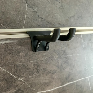 Può includere: Doppio gancio in metallo nero montato su una guida argentata contro un muro grigio con motivo marmoreo. I ganci sono curvi e progettati per appendere oggetti. Il muro ha un aspetto di pietra naturale.