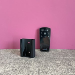 Op de afbeelding: Een zwarte wandhouder voor een afstandsbediening. De houder heeft twee gaten voor bevestigingsschroeven. Een zwarte afstandsbediening met het ZENEC-logo staat naast de houder.