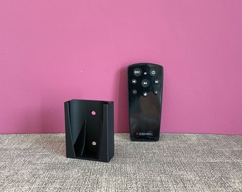 May include: A black wall mount holder for a remote control. The holder has two holes for mounting screws. A black remote control with a ZENEC logo is next to the holder.