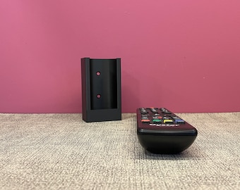 May include: A black plastic remote control holder with two holes for mounting on a wall. The holder is sitting on a grey textured surface. A black remote control with the word "Oyster" printed on it is sitting on the surface.