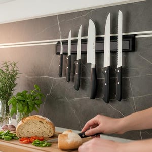 Könnte beinhalten: Ein Set Küchenmesser mit schwarzen Griffen und silbernen Klingen ist an einer Magnetleiste befestigt. Die Messer werden vor einer dunkelgrauen, marmorierten Rückwand präsentiert. Ein Holzschneidebrett mit Brot und Gemüse befindet sich im Vordergrund.