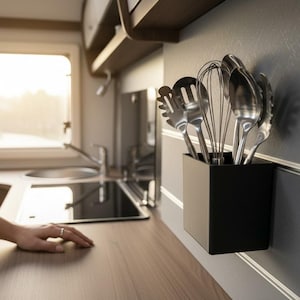 Könnte beinhalten: Ein schwarzer quadratischer Utensilienhalter an einer grauen Wand, gefüllt mit Küchenutensilien aus Edelstahl. Die Küche umfasst ein Waschbecken, ein Fenster und eine Holz-Arbeitsplatte. Eine Hand ruht auf der Arbeitsplatte.