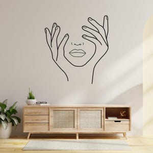 Peut inclure: Un dessin au trait minimaliste d'un visage encadré par des mains est monté sur un mur neutre. En dessous, un meuble en bois clair avec des panneaux de porte tissés et une plante en pot complètent le décor de la pièce. Un lampadaire avec un abat-jour beige se trouve à proximité.