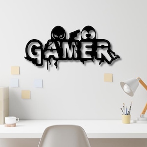 Peut inclure: Une enseigne murale noire représentant le mot GAMER dans une police stylisée. Le design comprend deux icônes de personnages circulaires avec des yeux positionnés au-dessus des lettres, et le bas des lettres présente un effet de peinture dégoulinante.