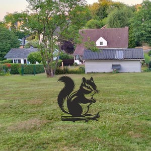 May include: A black metal silhouette of a squirrel perched on a branch, positioned in the center of a grassy lawn. The squirrel is depicted in profile with a bushy tail and small paws held near its chest, set against a background of houses.