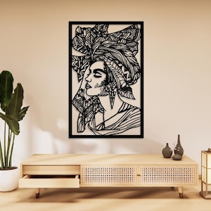 Peut inclure: Une décoration murale rectangulaire en métal noir représentant la silhouette découpée au laser d'une personne de profil, portant un foulard à motifs élaborés et de grandes boucles d'oreilles, dans un cadre noir.