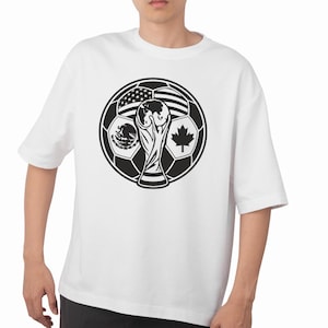 Può includere: Una t-shirt bianca a maniche corte con una grande grafica nera di un pallone da calcio. Il design del pallone incorpora le bandiere degli Stati Uniti, del Messico e del Canada nei suoi pannelli, centrato attorno a un'illustrazione di un trofeo.