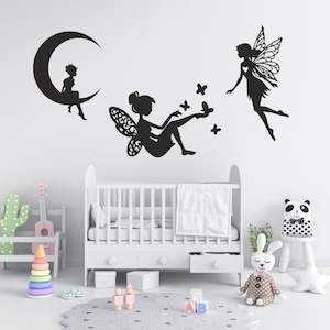 Peut inclure: Un ensemble d'autocollants muraux noirs représentant les silhouettes de trois fées et d'un croissant de lune. Une fée est assise sur la lune, une autre est assise avec des papillons et la troisième est en vol. Les motifs sont des formes noires.