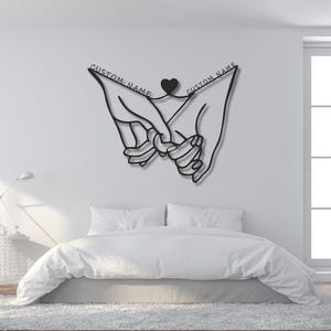Peut inclure: Une décoration murale en métal noir représentant un dessin au trait continu de deux mains aux doigts entrelacés. Un petit cœur est placé entre les poignets et le texte CUSTOM NAME est inscrit au-dessus de chaque main sur la structure.