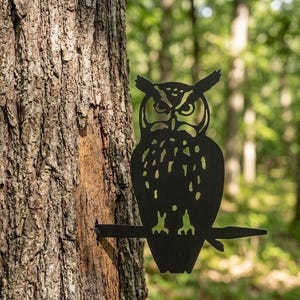Peut inclure: Une silhouette en métal noir représentant un hibou perché sur une branche. Le hibou présente des découpes détaillées pour les yeux, les traits du visage et les plumes de la poitrine. La figure est fixée au tronc d'un arbre par un piquet.
