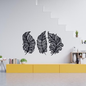 Peut inclure: Trois décorations murales en forme de plumes noires avec des motifs complexes. Les plumes sont montées sur un mur blanc au-dessus d'un meuble jaune. Les plumes présentent des motifs détaillés et des formes variées.