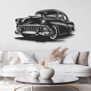 Peut inclure: Une décoration murale en noir et blanc représentant une voiture de style classique des années 1950 avec une calandre proéminente, des phares ronds et des pneus à flancs blancs. L'illustration présente des contrastes marqués sur un mur.