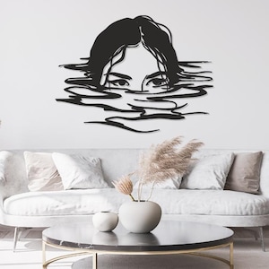 Peut inclure: Une décoration murale en métal noir représentant le haut du visage et les cheveux d'une personne émergeant de l'eau, avec des ondulations stylisées s'étendant horizontalement. Le design présente des lignes nettes et une finition mate.