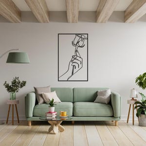 May include: Black metal wall art featuring a minimalist line drawing of a hand holding a rose. The artwork is framed in a rectangular black border. The piece is displayed on a light-colored wall above a green sofa in a living room setting.