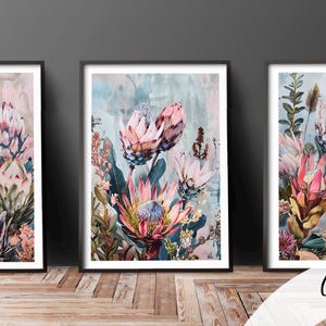 May include: Three framed floral art prints showcasing watercolour paintings of protea flowers in pink, peach, and blue hues. The artwork is displayed on a wooden surface against a grey wall.
