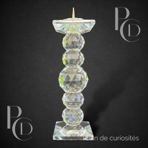 May include: A clear crystal candle holder with a square base and a tall, cylindrical column made of stacked crystal spheres. The holder has a small, gold-colored spike on top for holding a candle.