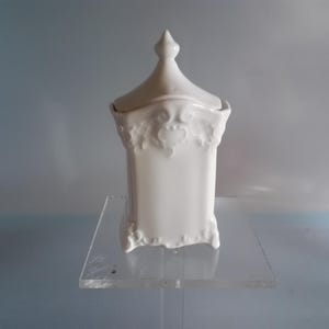 Puede incluir: Un tarro de cerámica blanco con tapa, con detalles ornamentales de relieve floral. El tarro tiene un cuerpo rectangular y un pináculo puntiagudo en la tapa. La pieza se exhibe sobre un soporte de acrílico transparente, sobre un fondo azul claro.