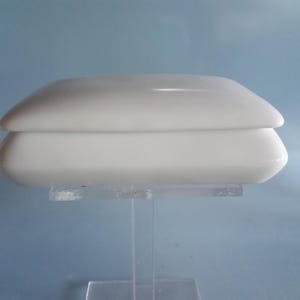 Op de afbeelding: Een witte, rechthoekige doos met deksel en afgeronde randen. De doos wordt gepresenteerd op een heldere acrylstandaard tegen een lichtblauwe achtergrond. De doos lijkt van keramiek of porselein te zijn.
