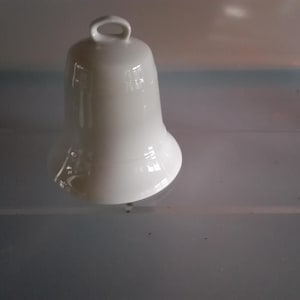 May include: A white ceramic bell with a loop at the top. The bell has a flared base and a smooth, glossy finish. The bell is a decorative item.