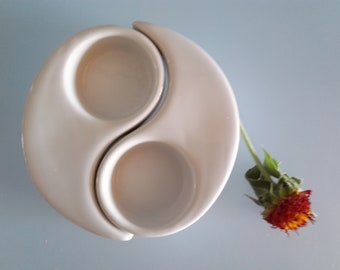 May include: White ceramic dish shaped like a yin yang symbol with two small bowls. The dish is on a light blue surface.