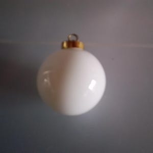 Puede incluir: Una bola de Navidad blanca y esférica con una tapa de metal dorada y un pequeño lazo para colgar. El adorno es liso y brillante, reflejando la luz. El fondo es gris apagado.