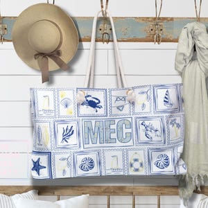 Op de afbeelding: Een strandtas met een blauw en wit ontwerp met illustraties van het zeeleven en de letters "MEC". Een strohoed en een lichtgekleurde sjaal hangen aan een verweerde blauwe houten plank met metalen haken. Twee kussens op een houten bank.