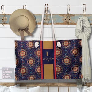 Puede incluir: Un bolso tote grande con un diseño azul marino y estampado, con una franja dorada central, la inicial "L" y el nombre "Lillian". El bolso cuelga de una pared con un sombrero y una bufanda. Cojines decorativos en un banco de madera.