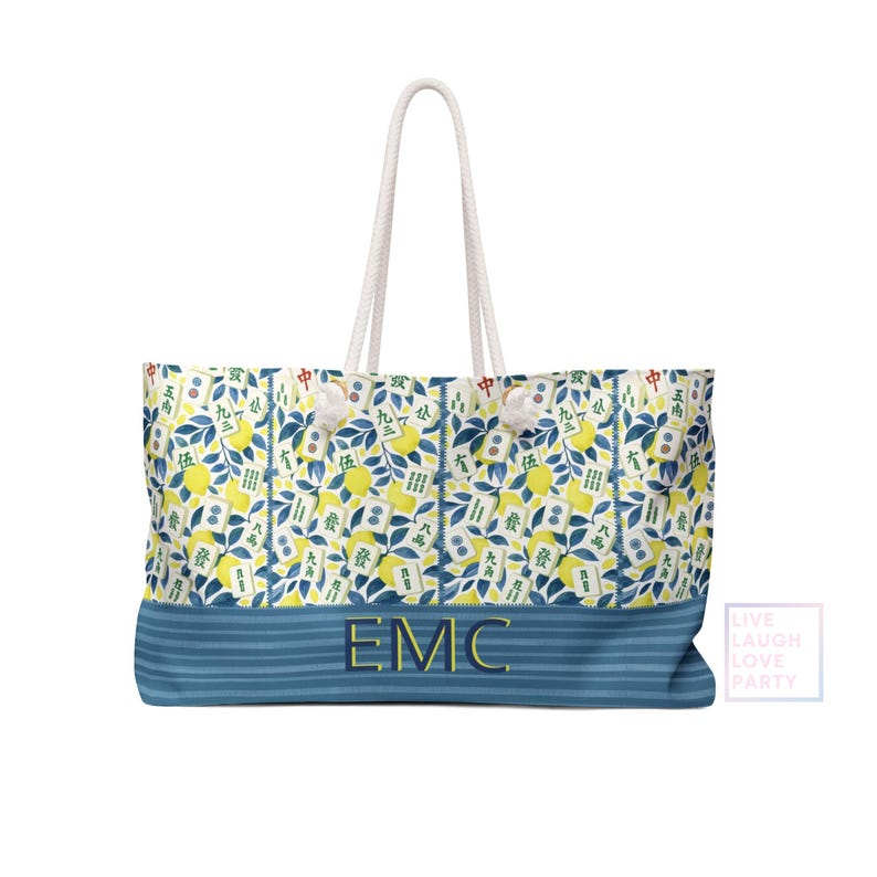 May include: A large tote bag with a white rope handle. The bag features a pattern of yellow lemons, blue leaves, and Chinese characters. The bottom of the bag is blue with the embroidered letters "EMC". The words "LIVE LAUGH LOVE PARTY" are in the corner.