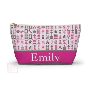 Puede incluir: Una bolsa rosa con cremallera y cremallera dorada, con un patrón de fichas de Mahjong. La sección inferior es de un fucsia vibrante con el nombre "Emily" en blanco. Una franja verde separa las dos secciones.