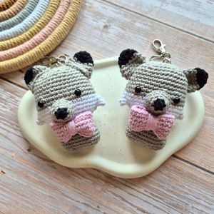 May include: Two grey crocheted fox keyrings with black eyes and pink bow ties. The keyrings are sitting on a white cloud-shaped surface.