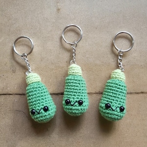 May include: Three handmade crochet keyrings shaped like green light bulbs with light green tops and black eyes. Each keyring has a silver metal ring and chain. The keyrings are displayed on a light brown surface.