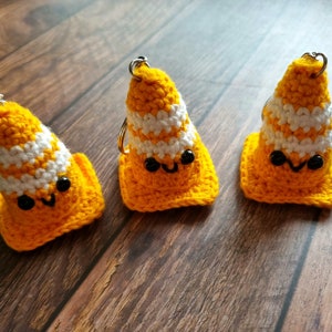 May include: Three handmade crochet keyrings shaped like orange and white traffic cones with smiling faces. Each keyring has a silver ring. The keyrings are on a wooden surface.
