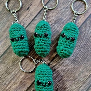 May include: Four green crocheted pickle keyring charms with black eyes and smiling faces. Each charm is attached to a silver key ring.