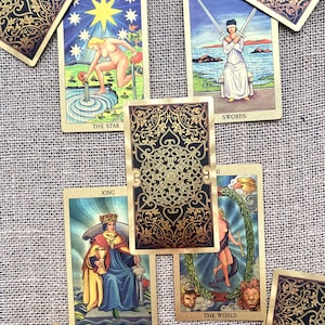 May include: A spread of tarot cards on a burlap background. The cards include The Star, Swords, King of Cups, and The World. The cards are decorated with gold accents and intricate designs.