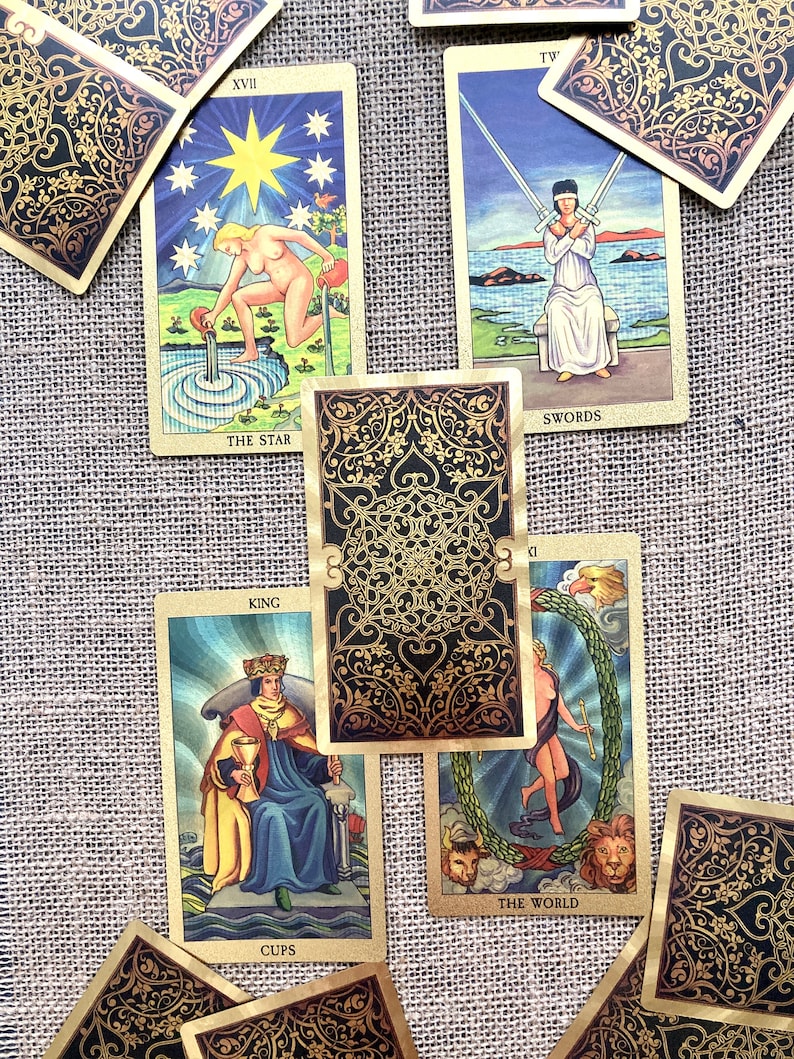 May include: A spread of tarot cards on a burlap background. The cards include The Star, Swords, King of Cups, and The World. The cards are decorated with gold accents and intricate designs.