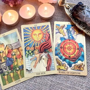 May include: Three tarot cards, The Sun, Wheel of Fortune, and Six of Cups, are laid out on a burlap surface. The cards are surrounded by three lit tea lights and a smudge stick on a gold metal holder.