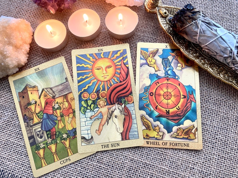 May include: Three tarot cards, The Sun, Wheel of Fortune, and Six of Cups, are laid out on a burlap surface. The cards are surrounded by three lit tea lights and a smudge stick on a gold metal holder.