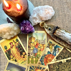 May include: A spread of tarot cards with gold accents and illustrations, including the Ace of Pentacles, Six of Cups, and the Sun card. A white sage smudge stick is in a gold holder, and there are three lit candles and crystals on a burlap background.
