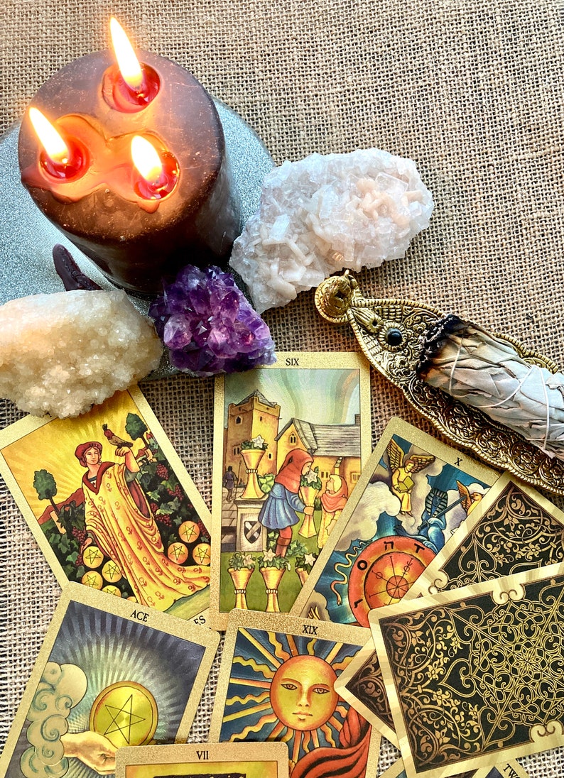 May include: A spread of tarot cards with gold accents and illustrations, including the Ace of Pentacles, Six of Cups, and the Sun card. A white sage smudge stick is in a gold holder, and there are three lit candles and crystals on a burlap background.