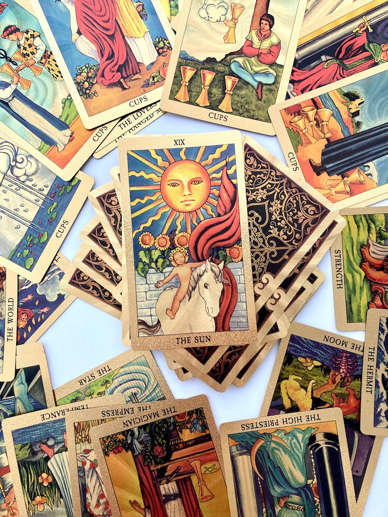 May include: A collection of tarot cards with gold accents and colorful illustrations. The cards are spread out on a white surface. The cards include The Sun, The World, The Hermit, The High Priestess, The Moon, Strength, The Lovers, The Magician, The Empress, The Star, and The Hanged Man.