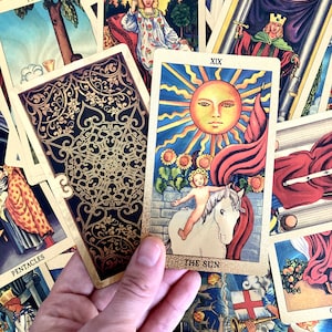 May include: The Sun tarot card, number XIX, depicts a nude child riding a white horse with a red flag behind them. The sun is shining brightly in the sky, and there are sunflowers in the background. The card is surrounded by other tarot cards.