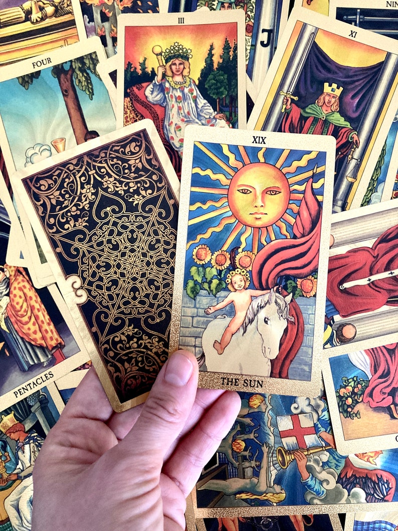 May include: The Sun tarot card, number XIX, depicts a nude child riding a white horse with a red flag behind them. The sun is shining brightly in the sky, and there are sunflowers in the background. The card is surrounded by other tarot cards.