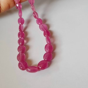 May include: A necklace of oval-shaped, polished pink gemstones. The beads are strung together, creating a continuous loop. The gemstones have a translucent quality, and the colour is a vibrant, saturated pink.