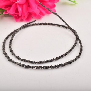 Puede incluir: Dos hilos de cuentas de diamantes en bruto, de color gris oscuro, dispuestas en forma circular sobre una superficie blanca. Una vibrante flor rosa y follaje verde son visibles en el fondo, a&ntilde;adiendo un toque de color. Las cuentas miden unos 40 cm de largo.