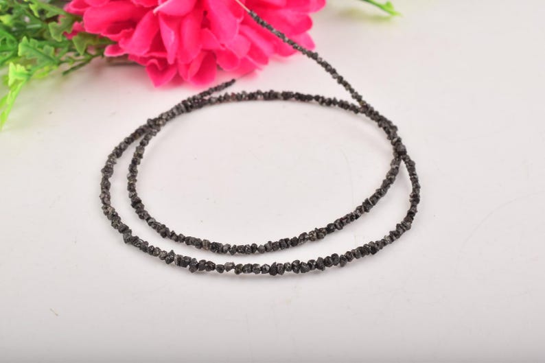 Puede incluir: Dos hilos de cuentas de diamantes en bruto, de color gris oscuro, dispuestas en forma circular sobre una superficie blanca. Una vibrante flor rosa y follaje verde son visibles en el fondo, a&ntilde;adiendo un toque de color. Las cuentas miden unos 40 cm de largo.