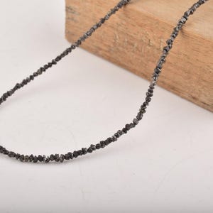 Puede incluir: Un collar de cuentas negras compuesto por peque&ntilde;as cuentas de forma irregular. El collar est&aacute; colocado sobre un bloque de madera, sobre un fondo blanco. Las cuentas parecen ser de color gris oscuro.